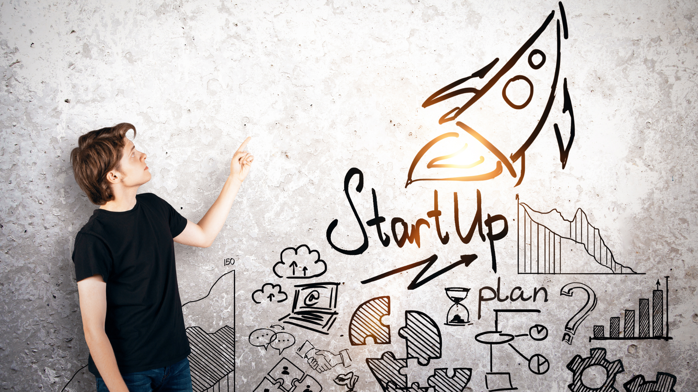 A young man pointing to a wall with symbols drawn by marker. It represents startups, with a upward rocket ship and puzzle pieces, communication, time, planning, website creation, and other things one would need to do.