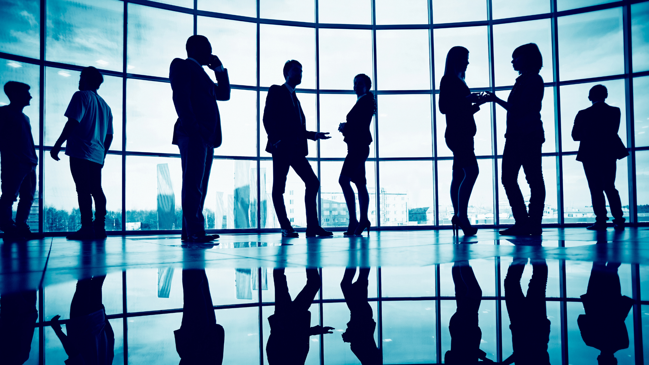 Untitled design (22) A large office window with a city view has seven professionally dressed individuals nearby either talking to each other or on their cellphone.