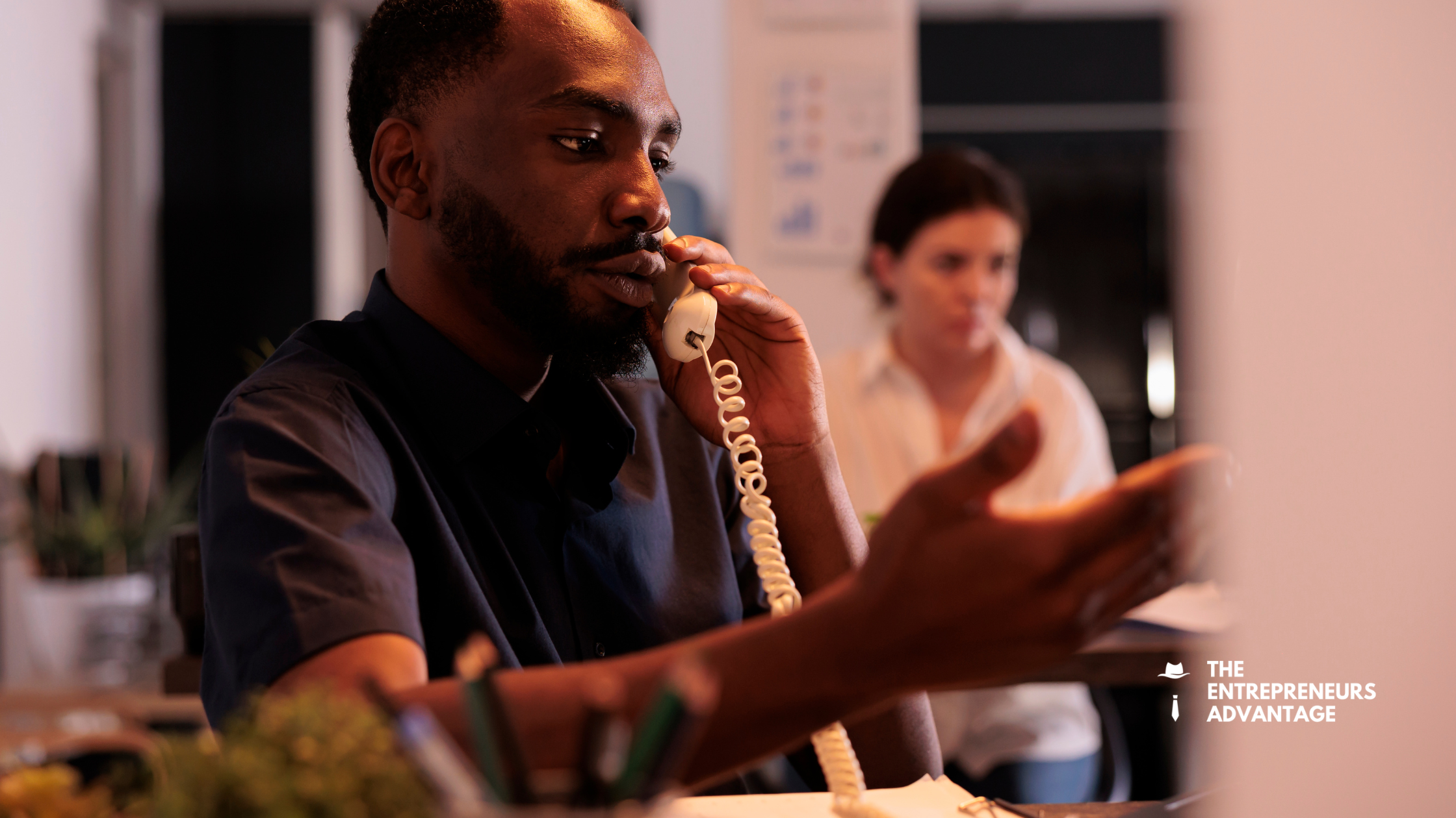A busy male entrepreneur talking on a desk phone while looking stressed in an office setting with a blurred coworker in the background.