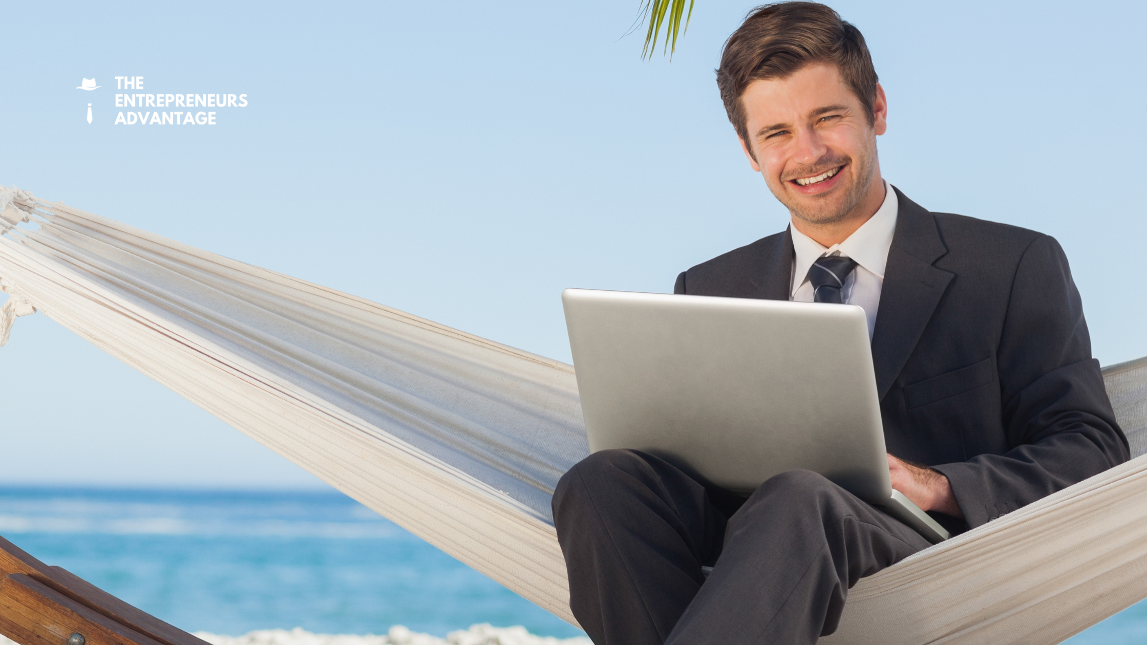 Summer Business Automation – Take a Vacation Without Business Catching Fire A smiling businessman in a suit relaxing in a hammock on a tropical beach while using a laptop, representing the freedom of taking a summer vacation when your business is fully automated.